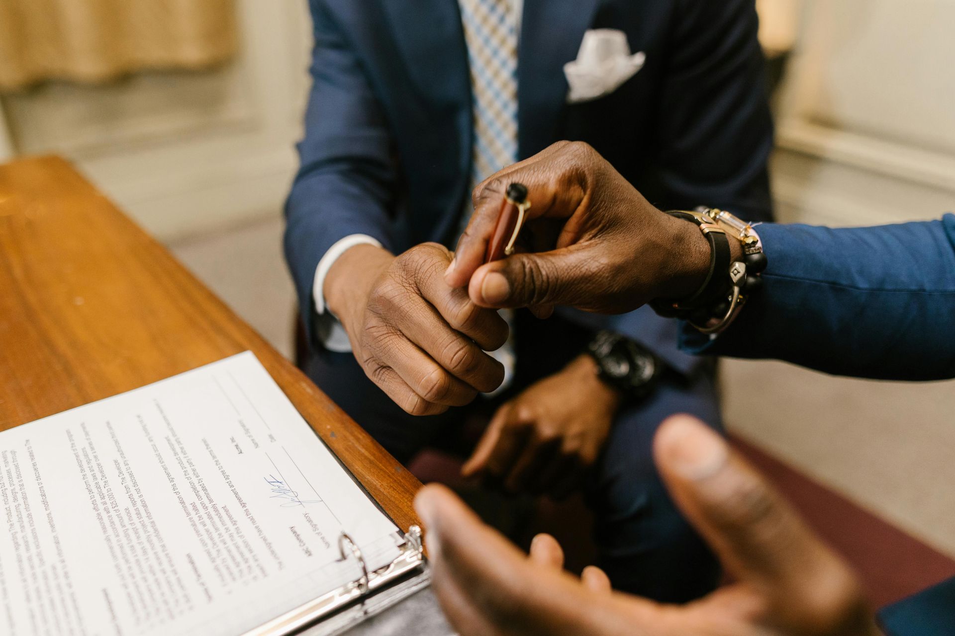 Hands passing a pen over a document on a table. One person wears a navy suit.