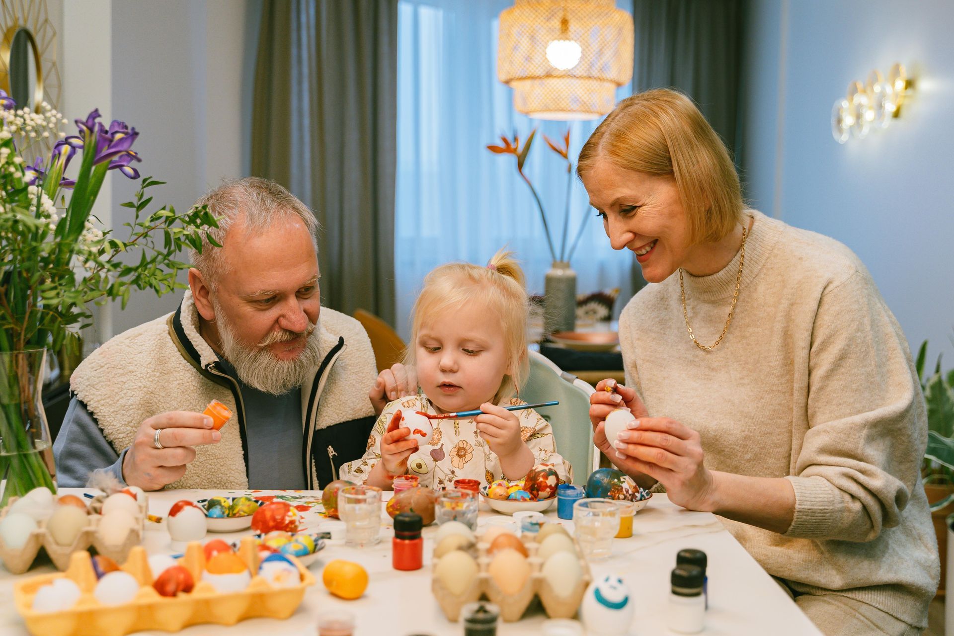 Family painting Easter eggs at a table. Child in the center, grandparents flanking her, all smiling.