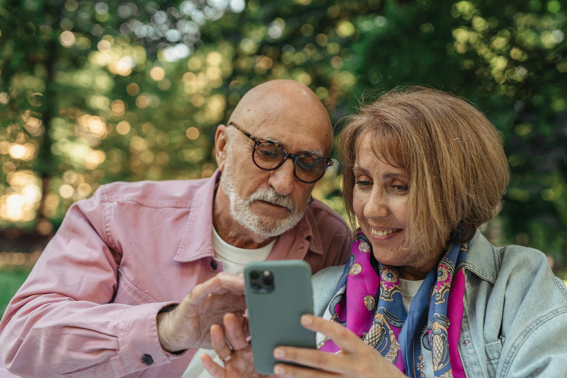 Older couple looking at a smartphone screen outdoors in a park. Man pointing; woman smiling.