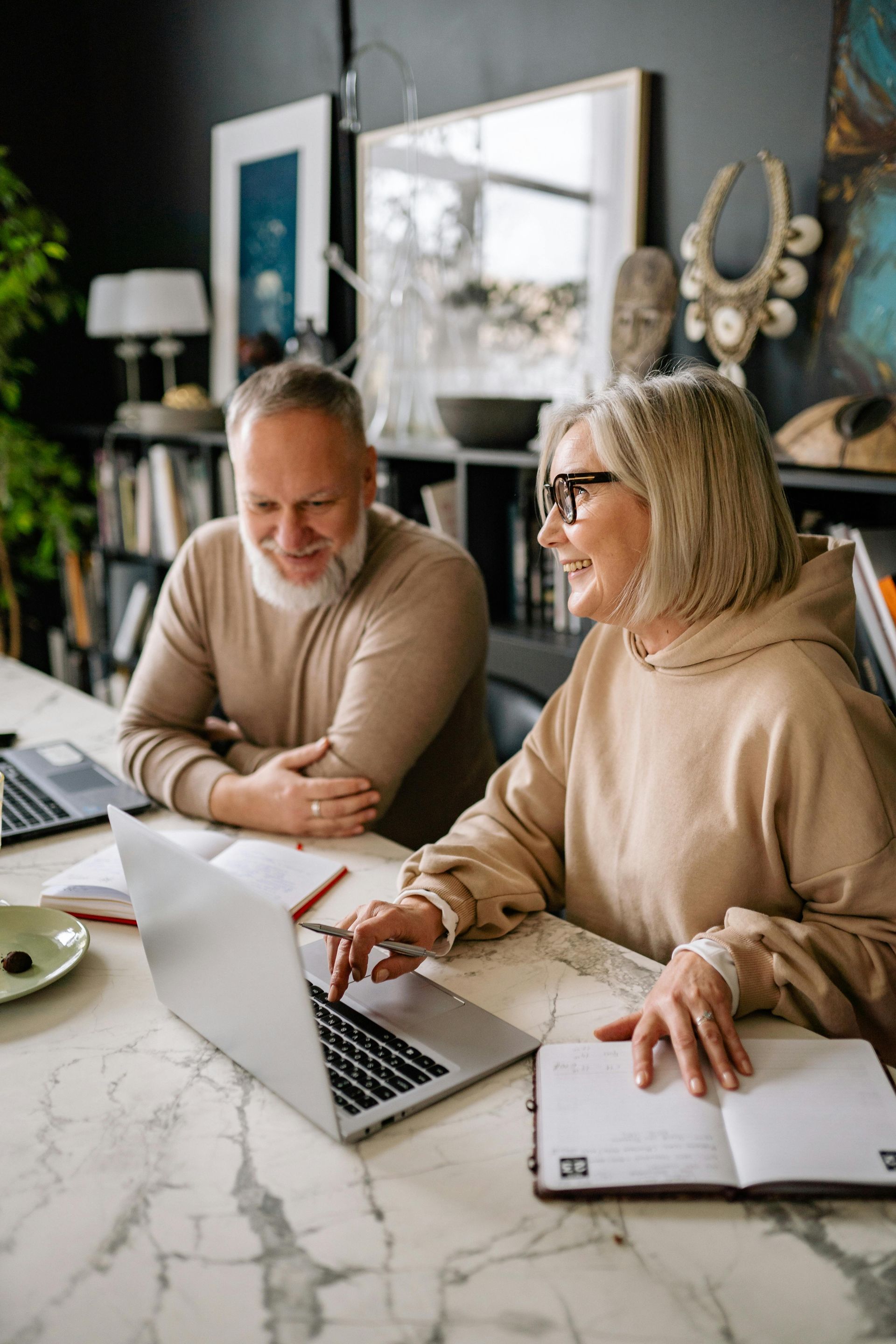 Older couple looking at a laptop together, smiling, at a white marble table. Bookshelves and art in the background.