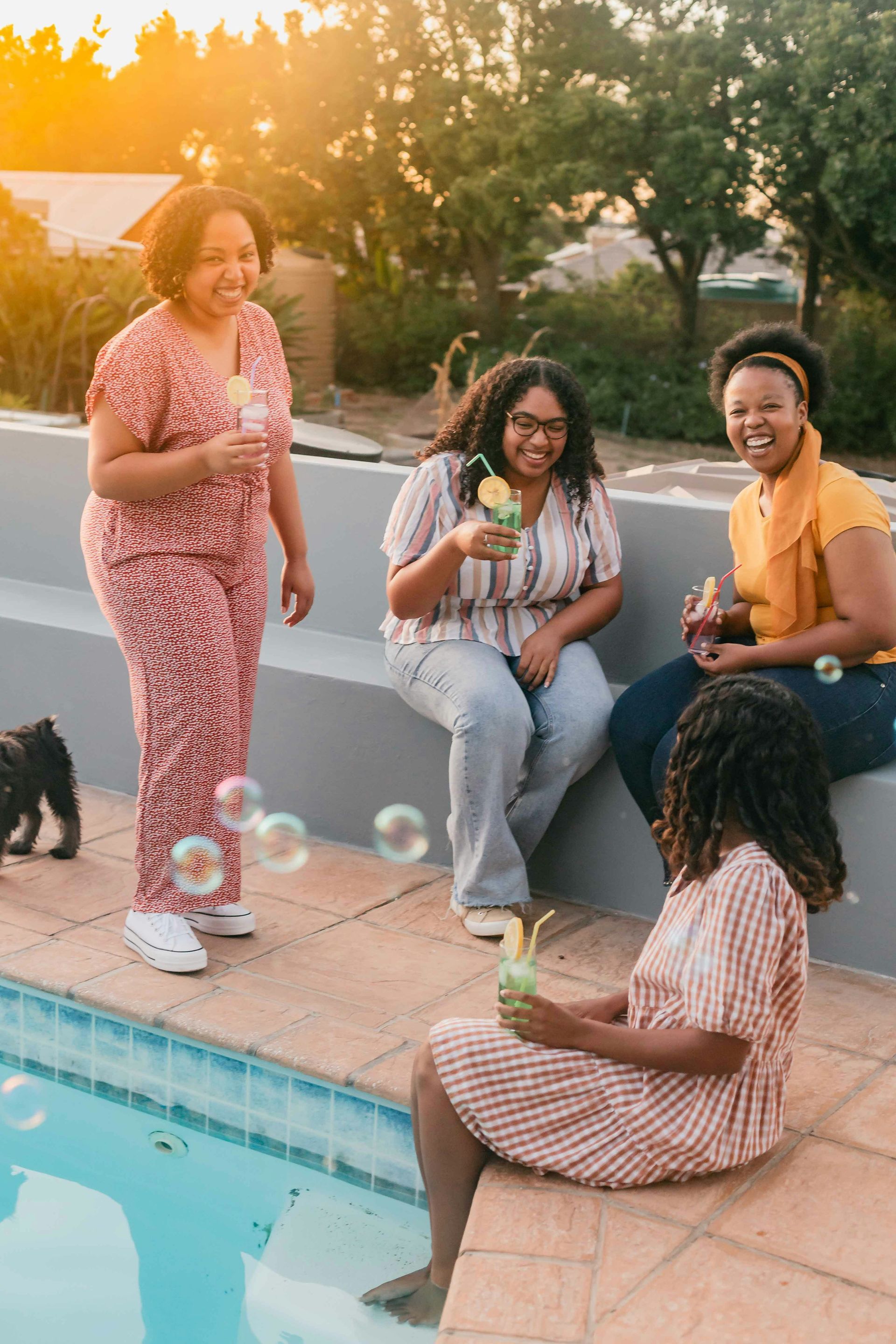 Four people around a pool, blowing bubbles. One sits, three stand and smile, golden hour lighting.