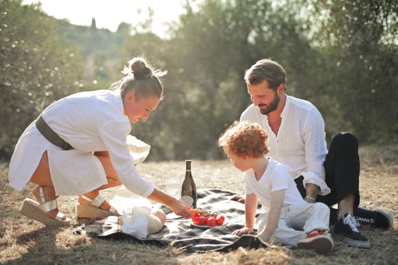 A family in white clothing has a picnic on a blanket in a sunny outdoor setting with trees in the background.