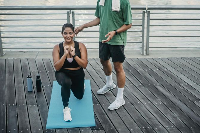 A person performs a lunge on a blue mat outdoors while a trainer stands nearby on a wooden deck.