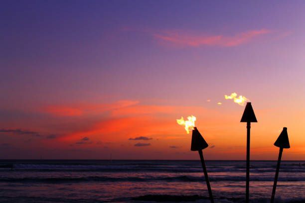 Sunset over ocean with tiki torches ablaze. Sky with orange and purple hues.