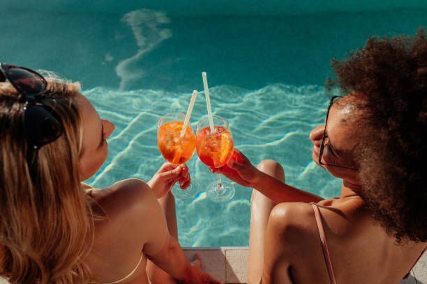 Two people toasting orange drinks by a pool.