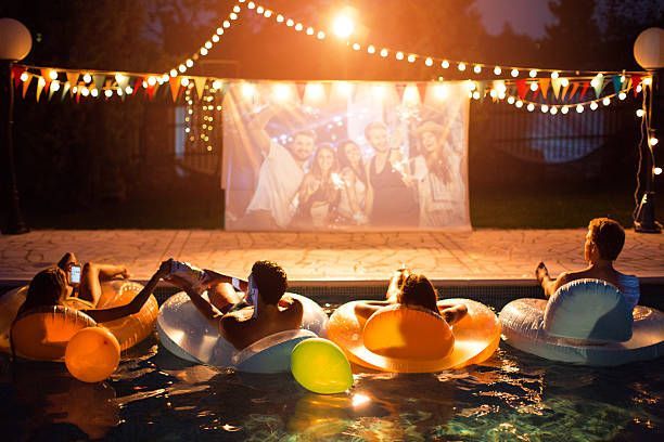 People in pool watching movie on screen; string lights, inflatable rings, and party decorations.