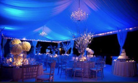 Blue-lit tent decorated for a formal event with tables, chairs, flowers, and chandeliers.