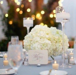Wedding table setting with white flowers, candles, and a table number against a blurred background of lights.