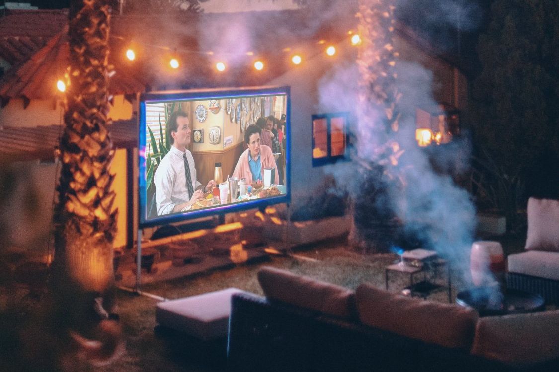 Outdoor movie night setup: large screen, fairy lights, seating, and a fire pit with smoke.