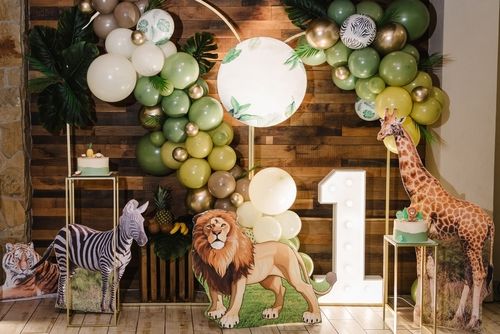 Jungle-themed first birthday party decorations: Balloons, animal cutouts, cake stands, lit-up number one, and a wooden backdrop.