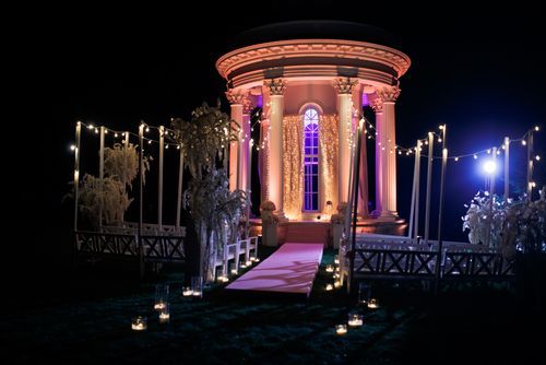 Nighttime view of a gazebo lit with pink and white lights, pathway with candles.