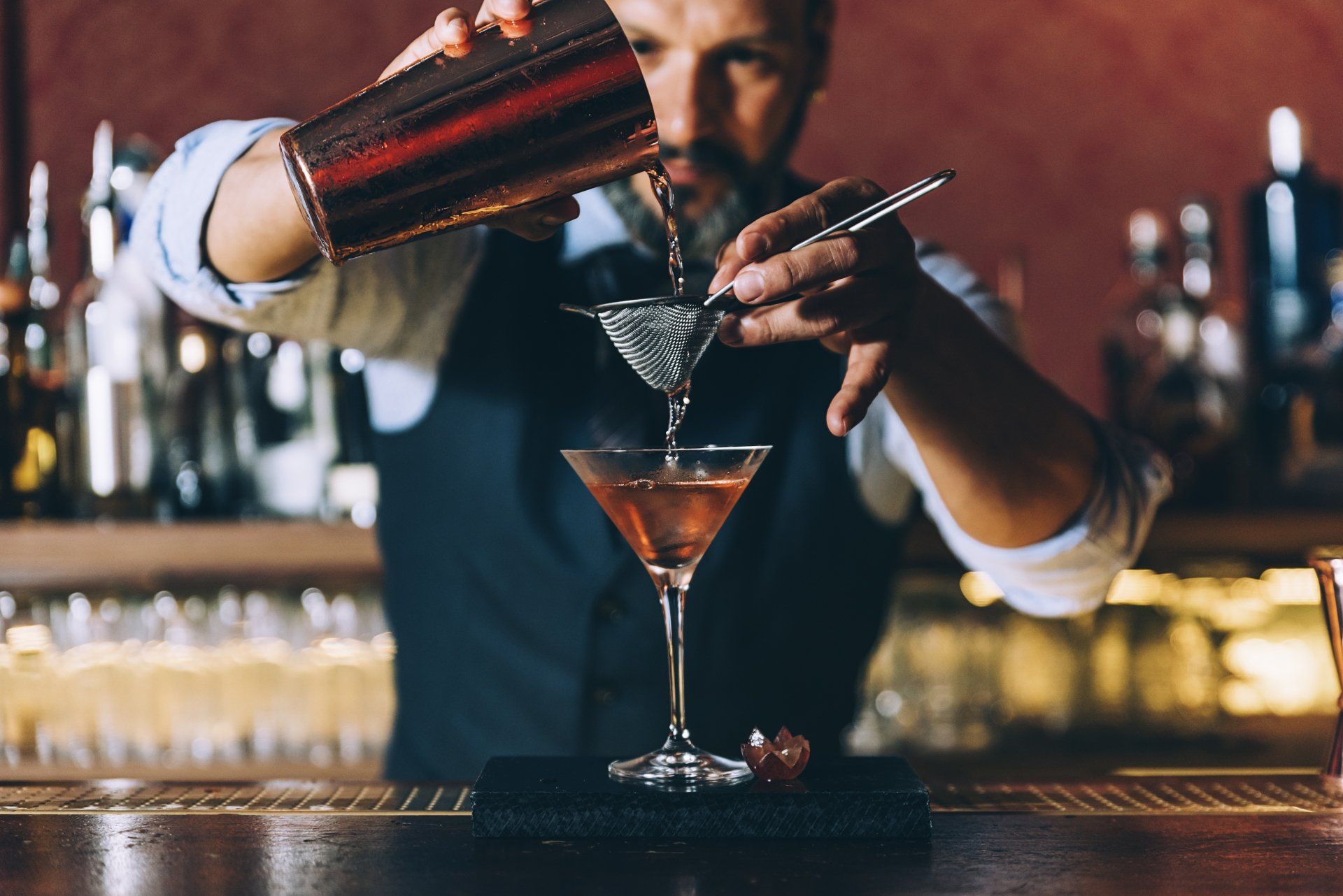Bartender pouring a drink from a shaker into a martini glass, bar setting.