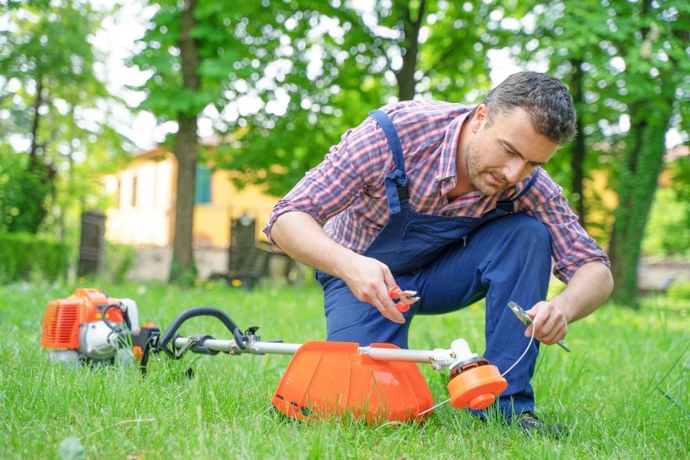 Man in blue overalls repairs a string trimmer on a lawn. Orange trimmer, green grass, trees in background.