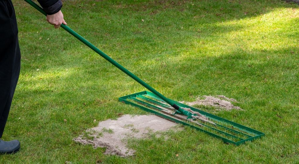 Person raking sand or soil on a green lawn with a long-handled leveler.