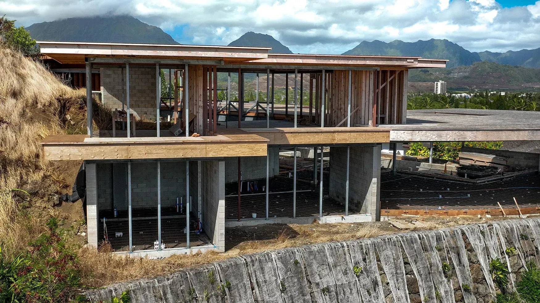 Unfinished modern house on a hillside with glass walls and wooden framing, mountain backdrop.