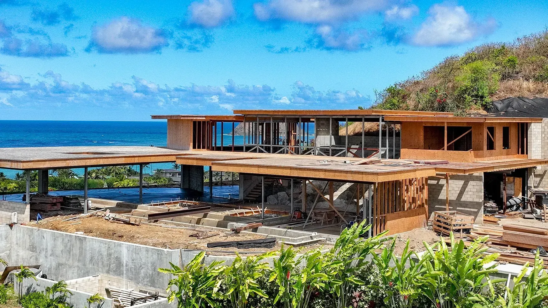 House under construction overlooking the ocean with blue sky, green foliage, and wooden framing.