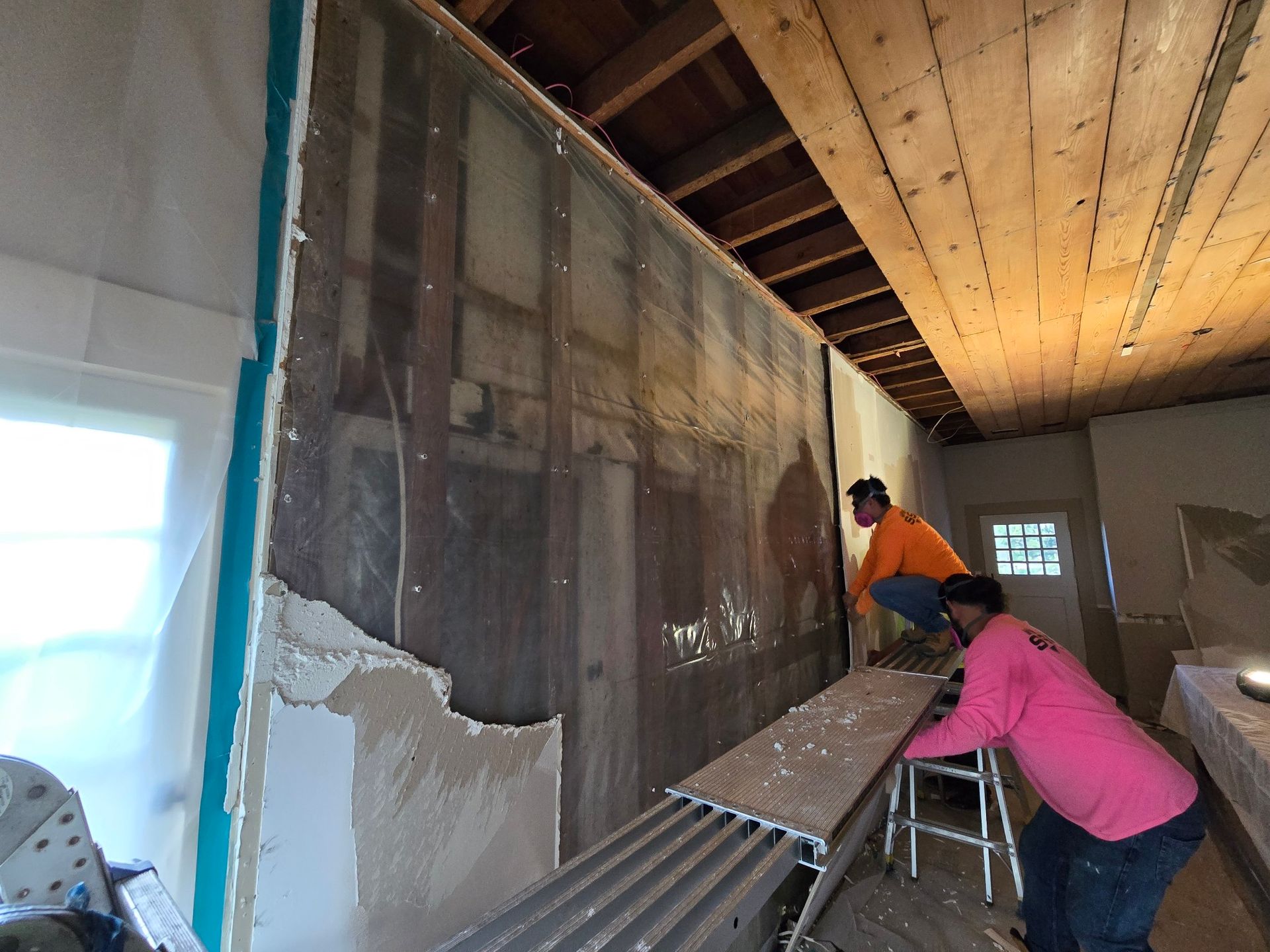 Workers removing wall material in a room with exposed ceiling beams. One stands on a ladder.