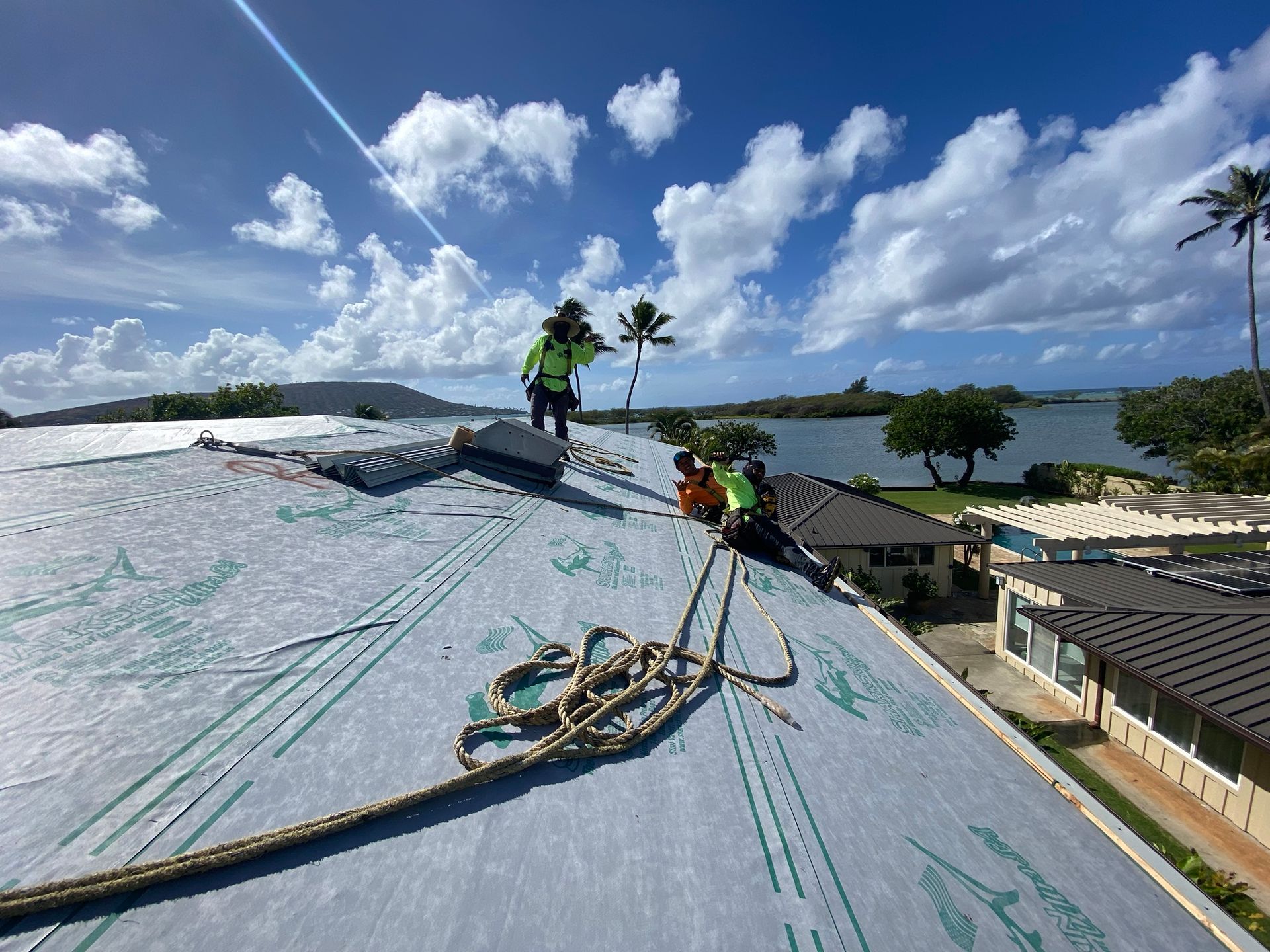 Roofers working on a residential roof near water with bright sky.