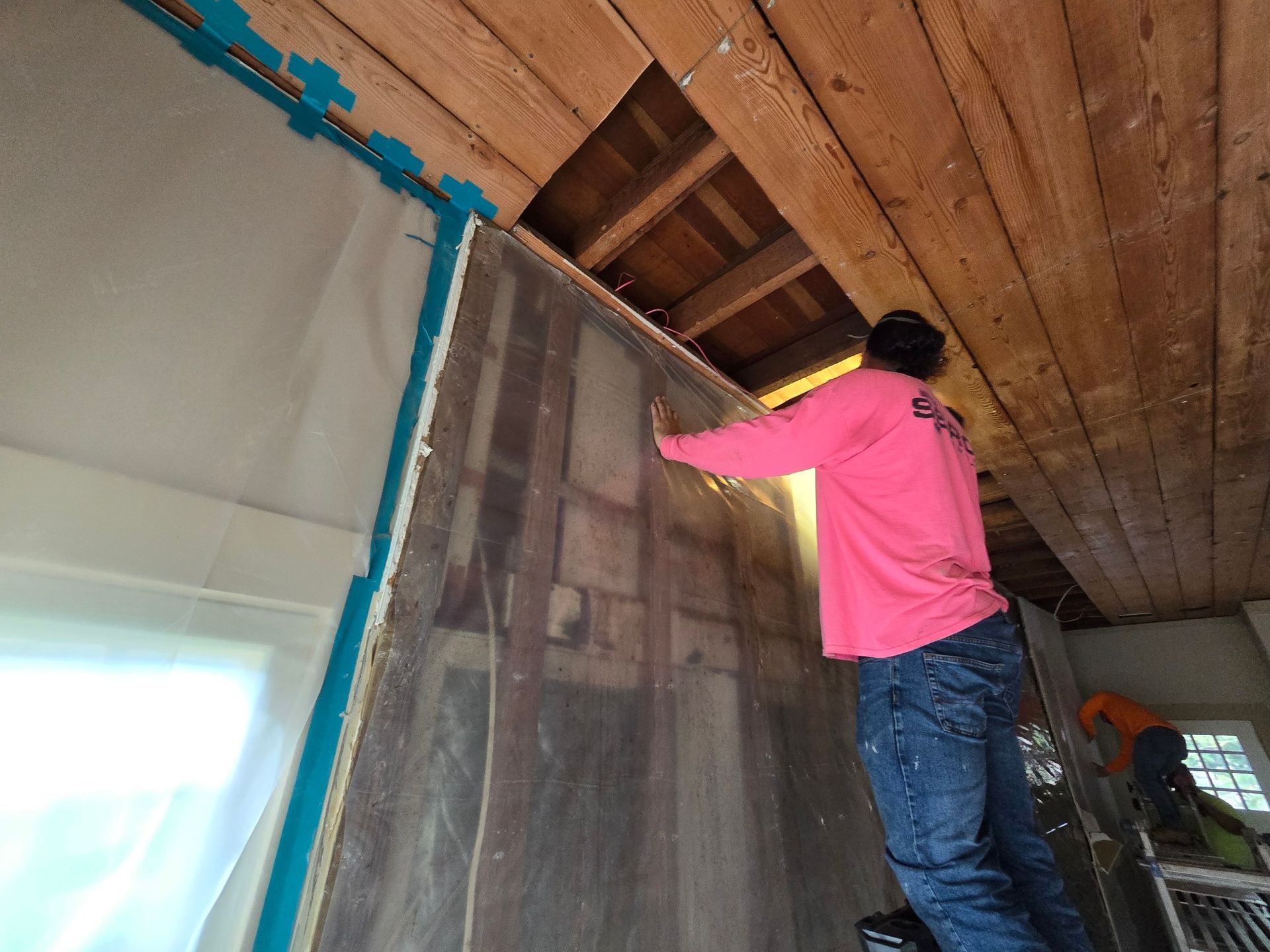 A person in a pink shirt is installing a wall panel indoors with exposed wooden beams overhead.