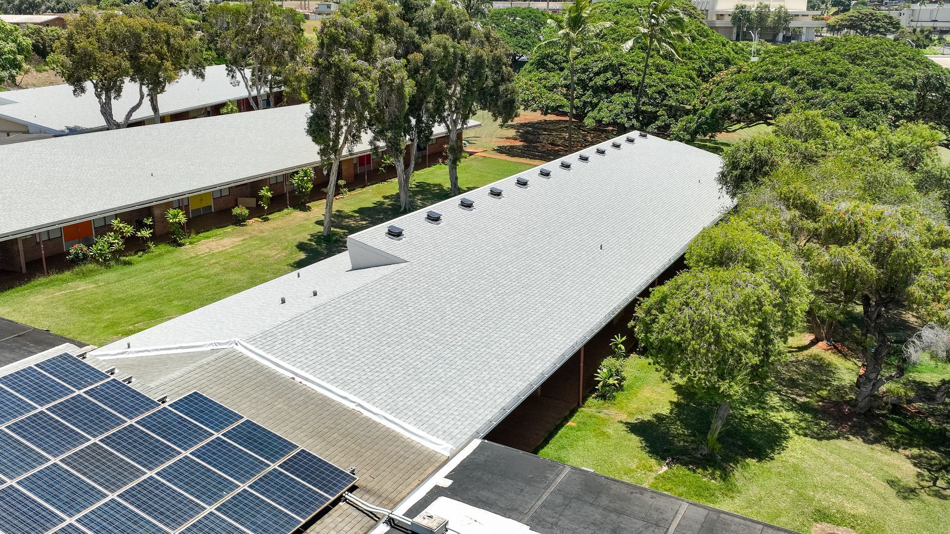 Overhead view of a long, gray building with corrugated metal roof and rows of small vents, surrounded by trees and green grass.