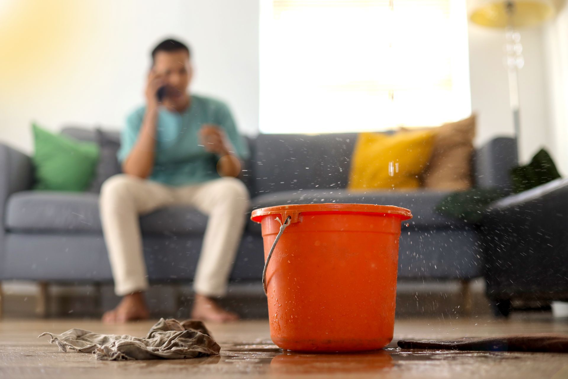 An orange bucket is catching water from a roof leak, with a man in the background making a call