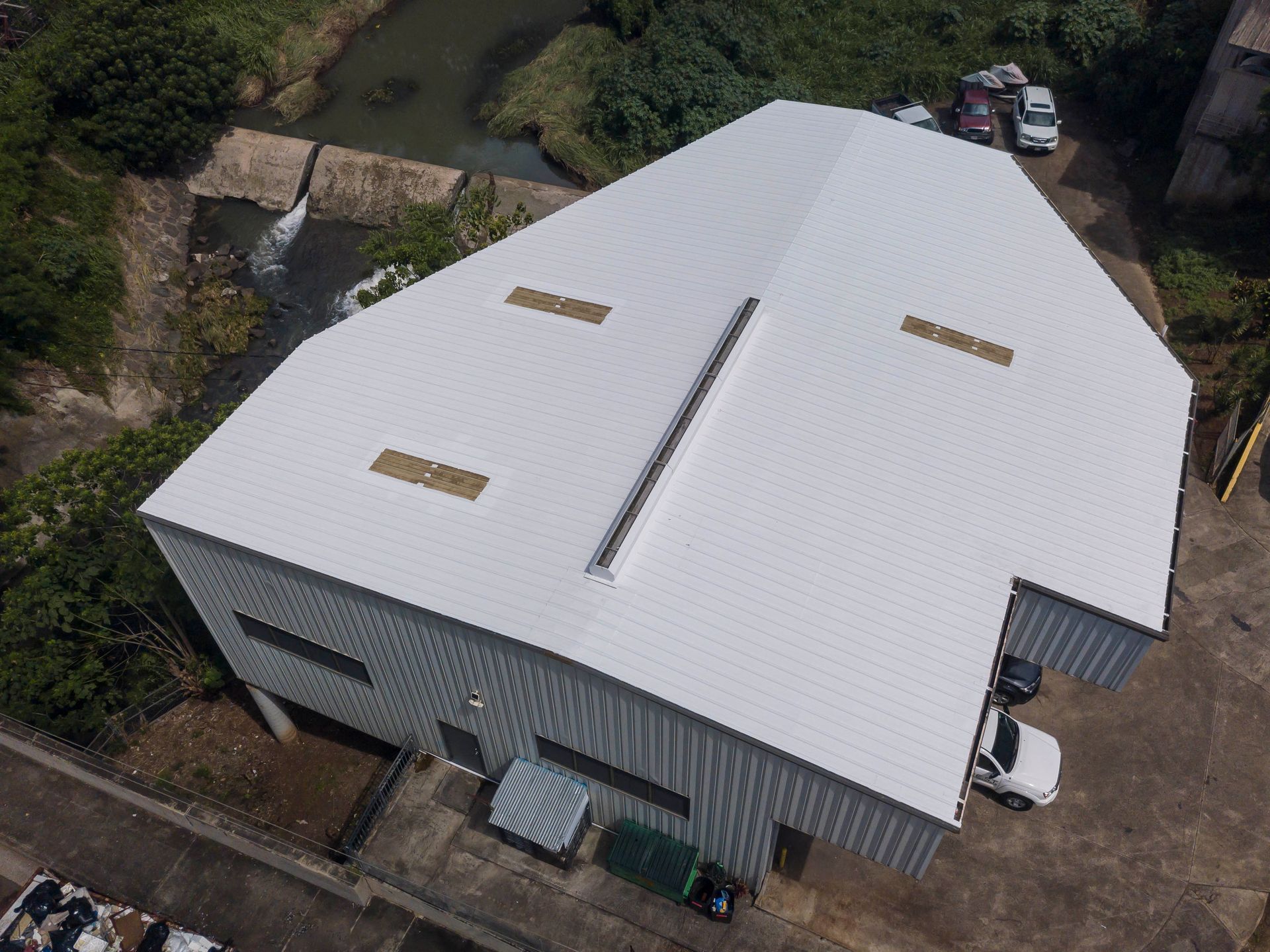 Overhead view of a white industrial building with a corrugated metal roof near a waterway and parked vehicles.
