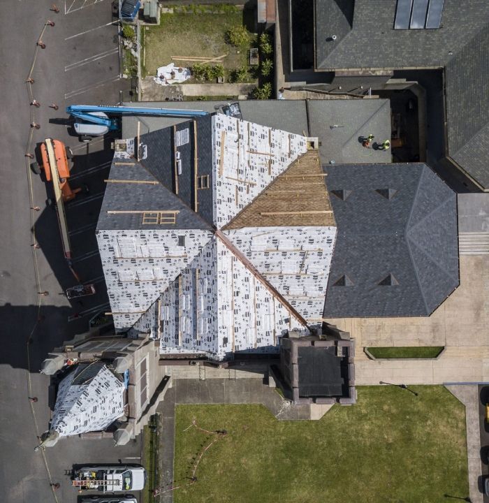 Aerial view of a building roof under construction. Gray and white roofing materials visible; workers on roof.