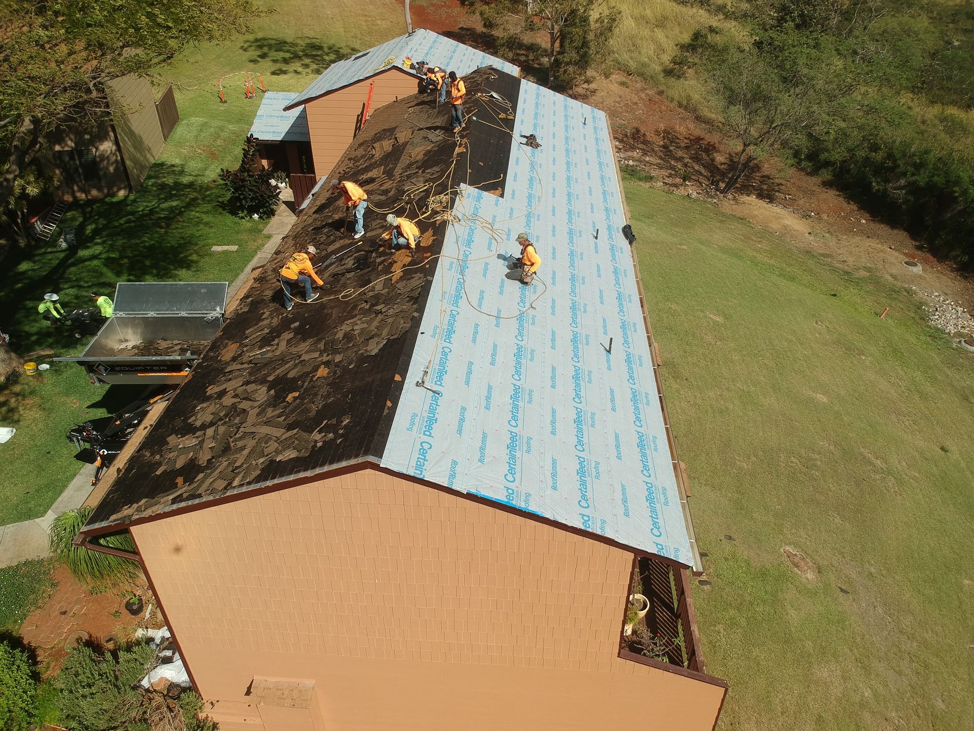 Roofers installing a new roof on a brown building; workers in yellow vests on a grassy hillside.
