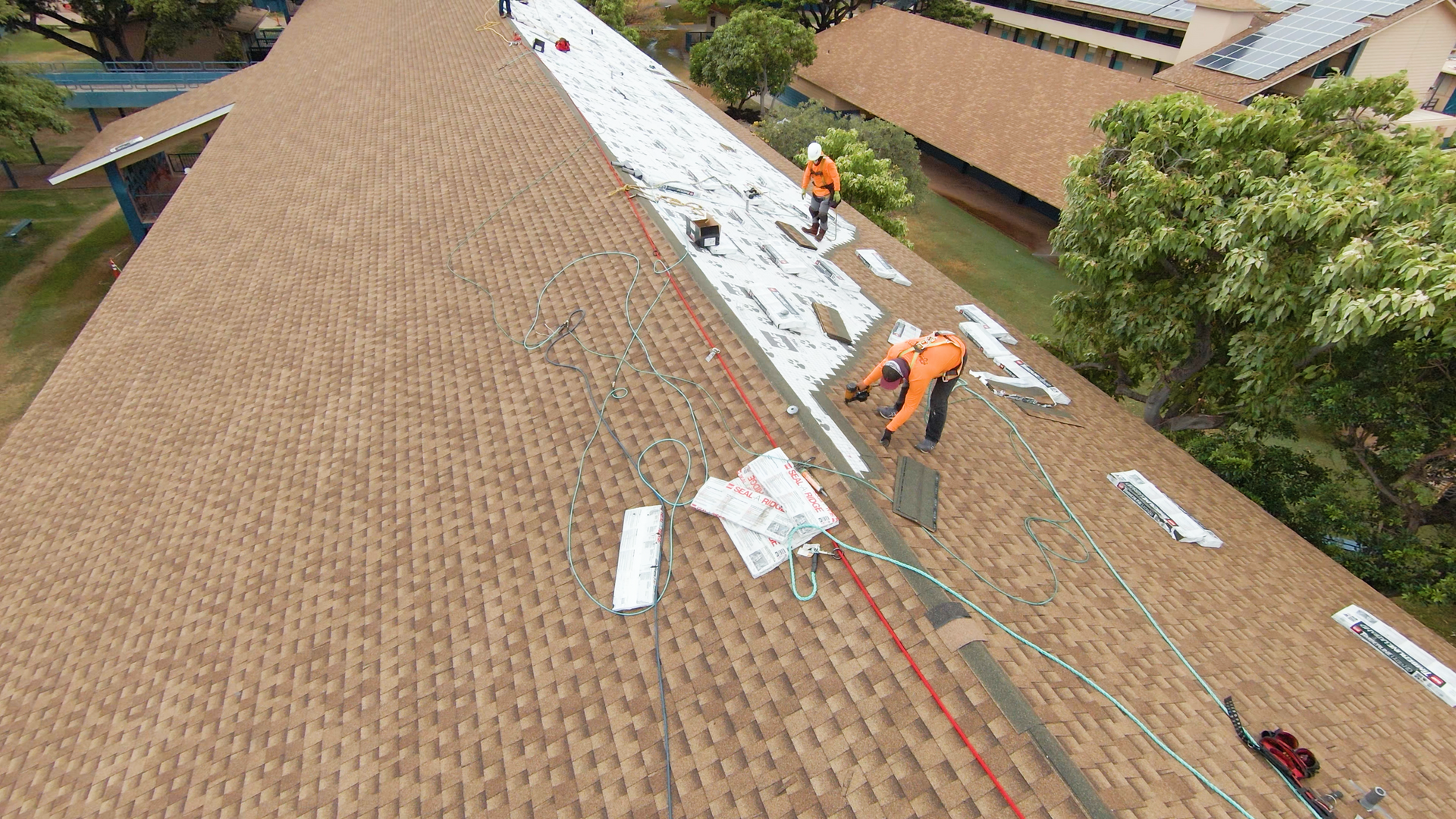 Workers in orange suits repairing a brown shingle roof, partially covered with white material.