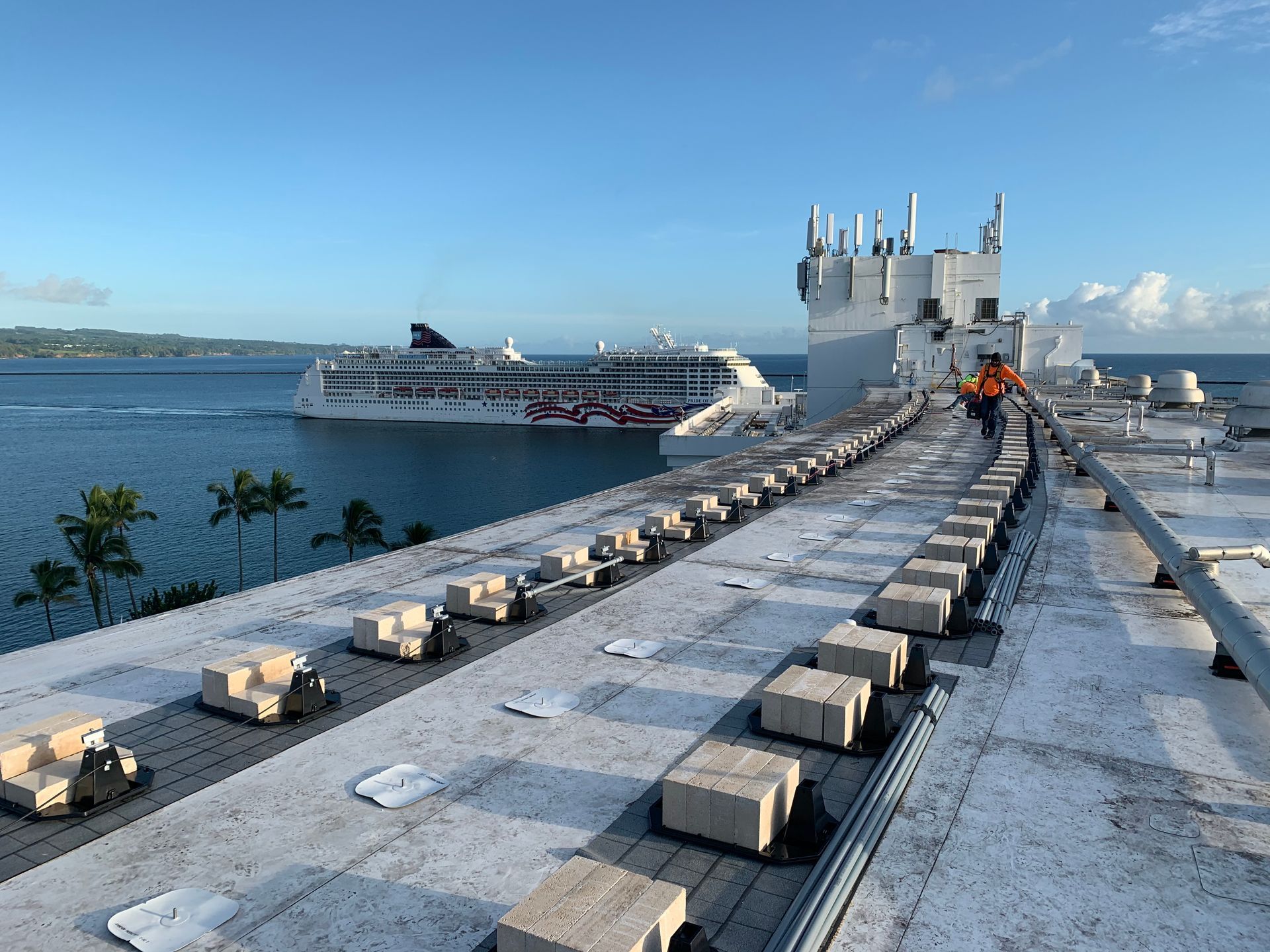 Rooftop with a worker, a cruise ship, and ocean view. Gray and white building with a sunny sky.