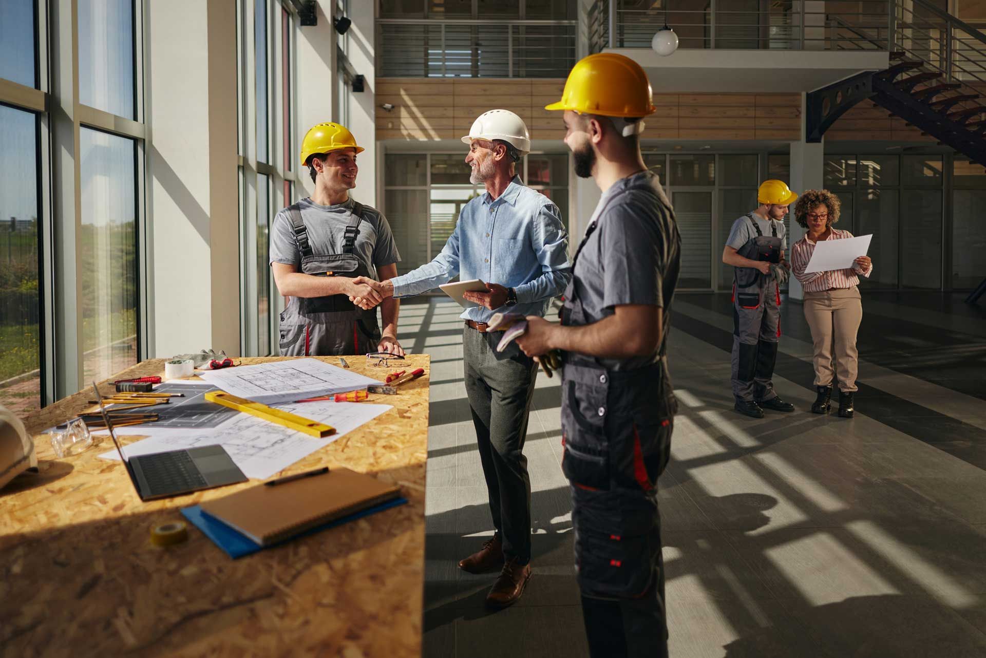 A happy project manager greeting commercial general contractors, in yellow helmets, in a building.