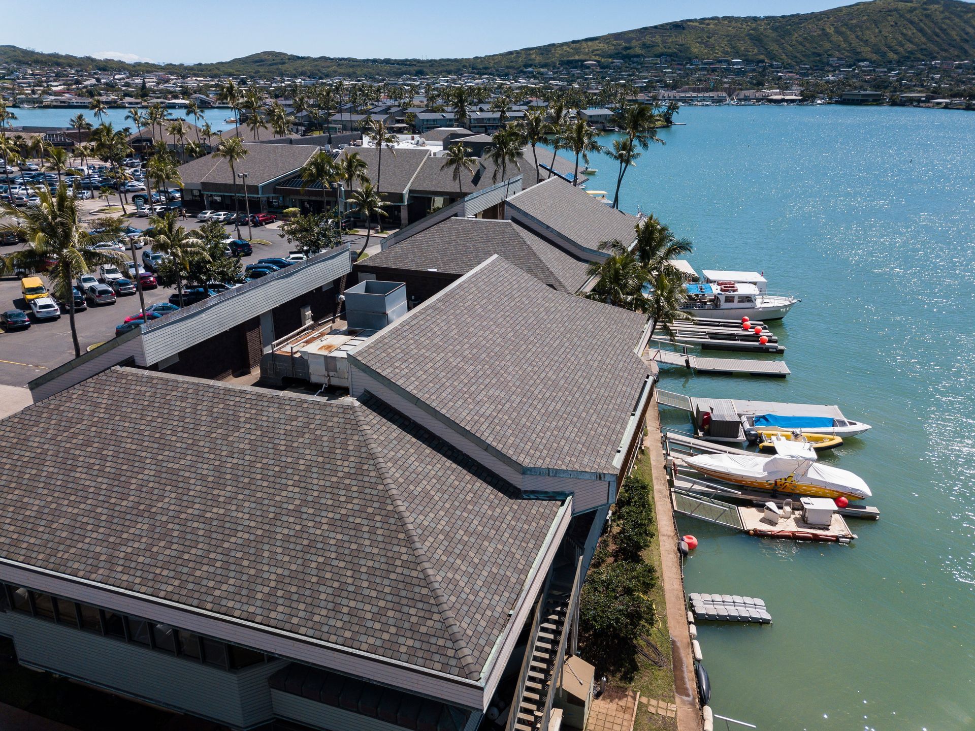 Buildings with gray roofs next to a harbor with boats; mountain backdrop, blue water.