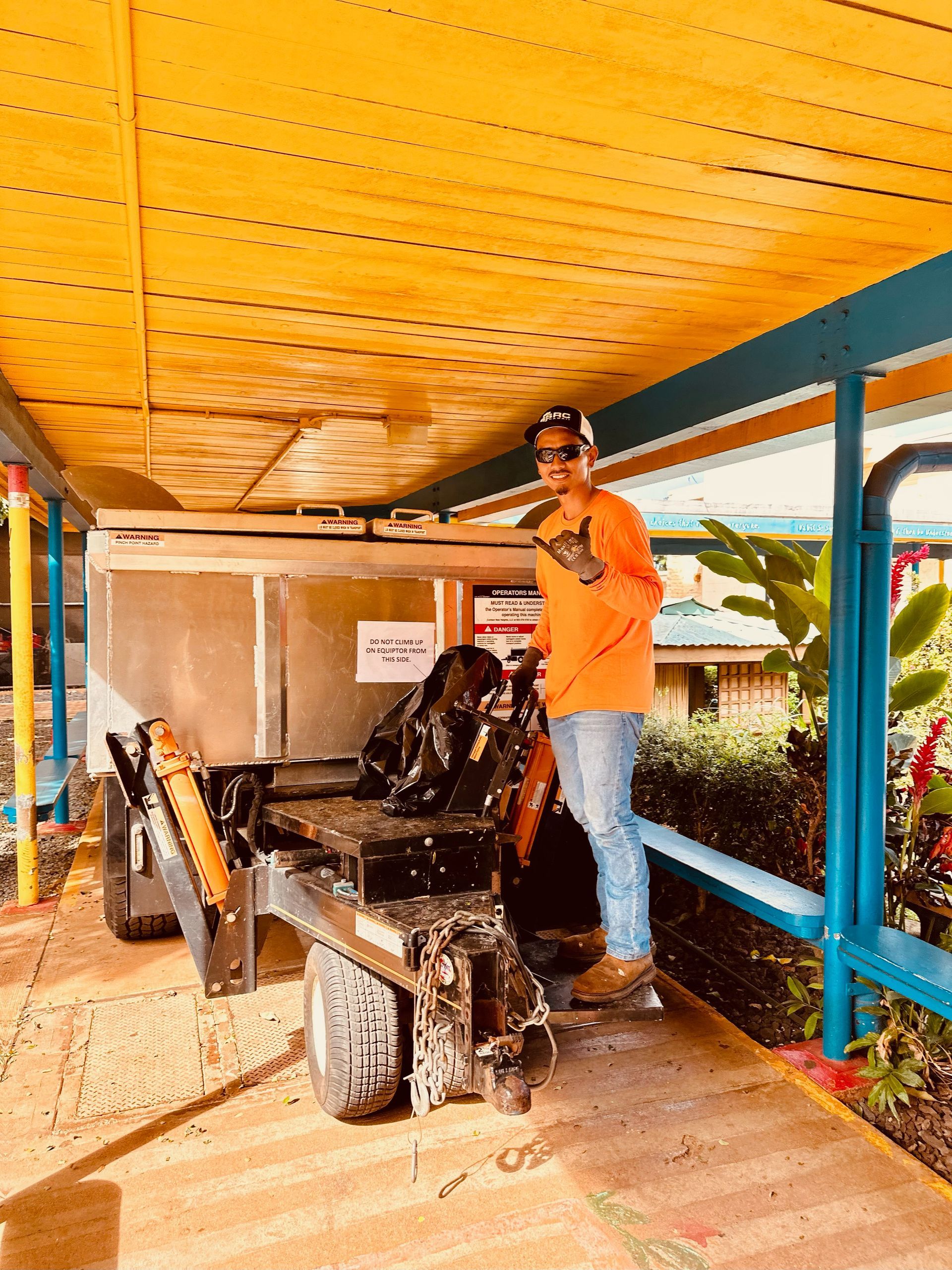Man in orange shirt standing next to a small trailer, in a covered walkway, blue beams, and a yellow ceiling.