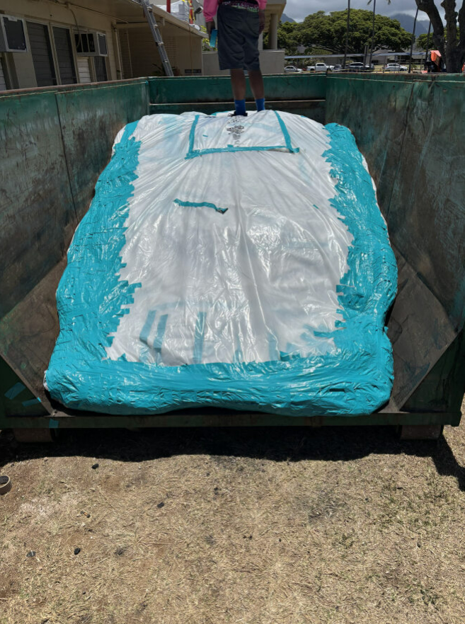 Person standing on a large, blue-edged, white tarp inside a green dumpster. Sunny outdoor setting.
