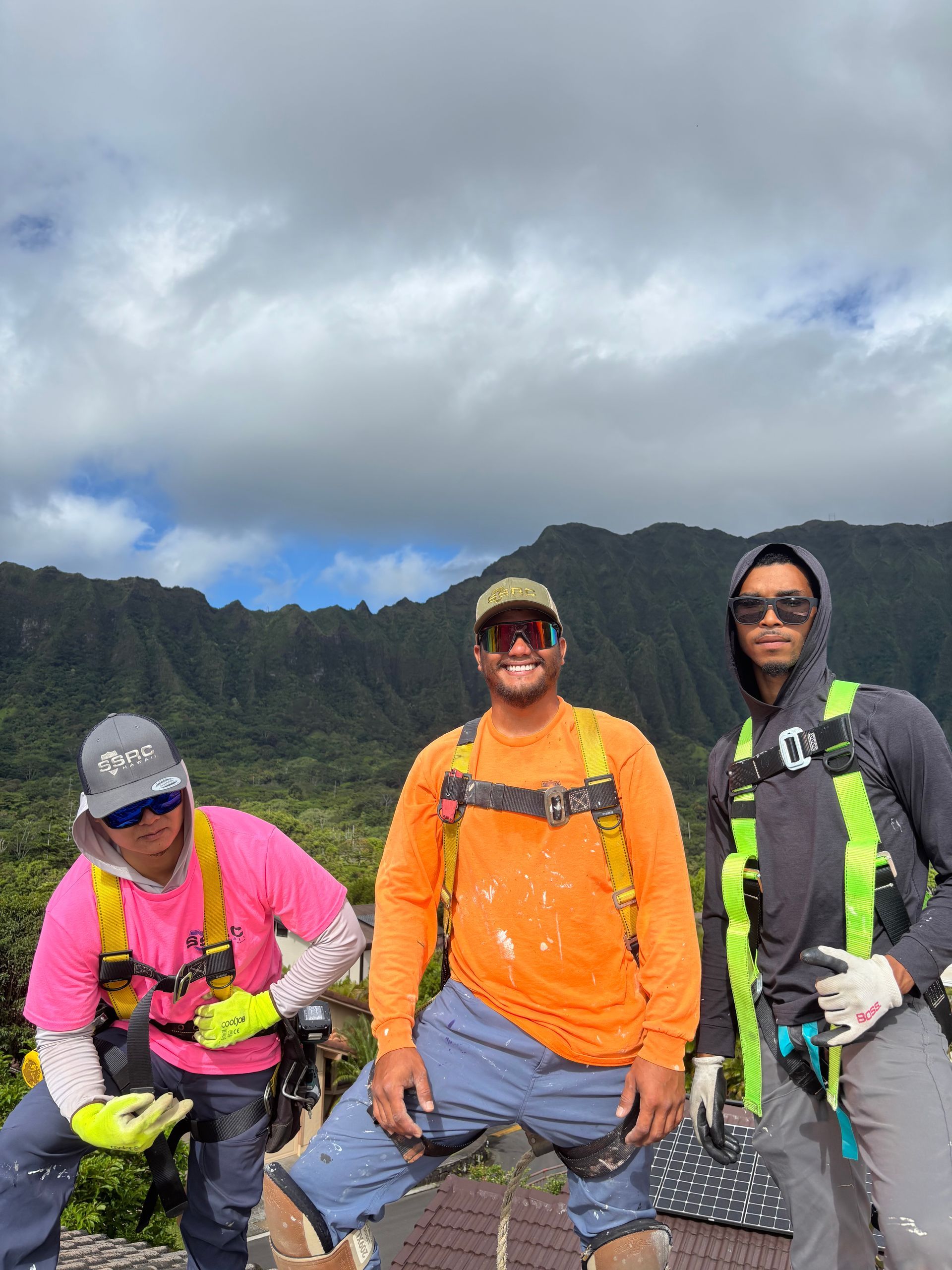 Three construction workers on a rooftop wearing safety harnesses, posing with a mountain background.
