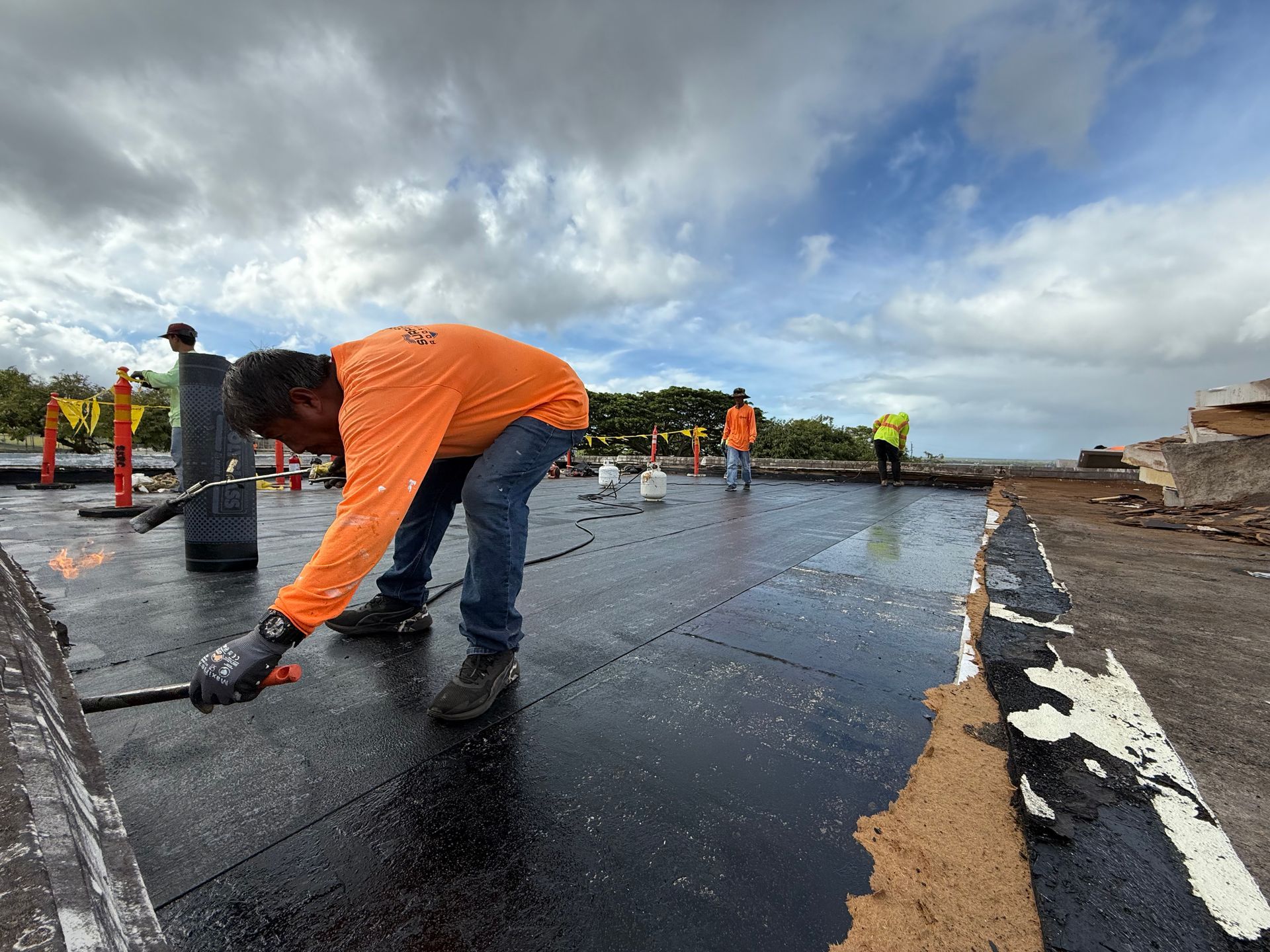 Construction worker in orange shirt torches asphalt roofing on a cloudy day.