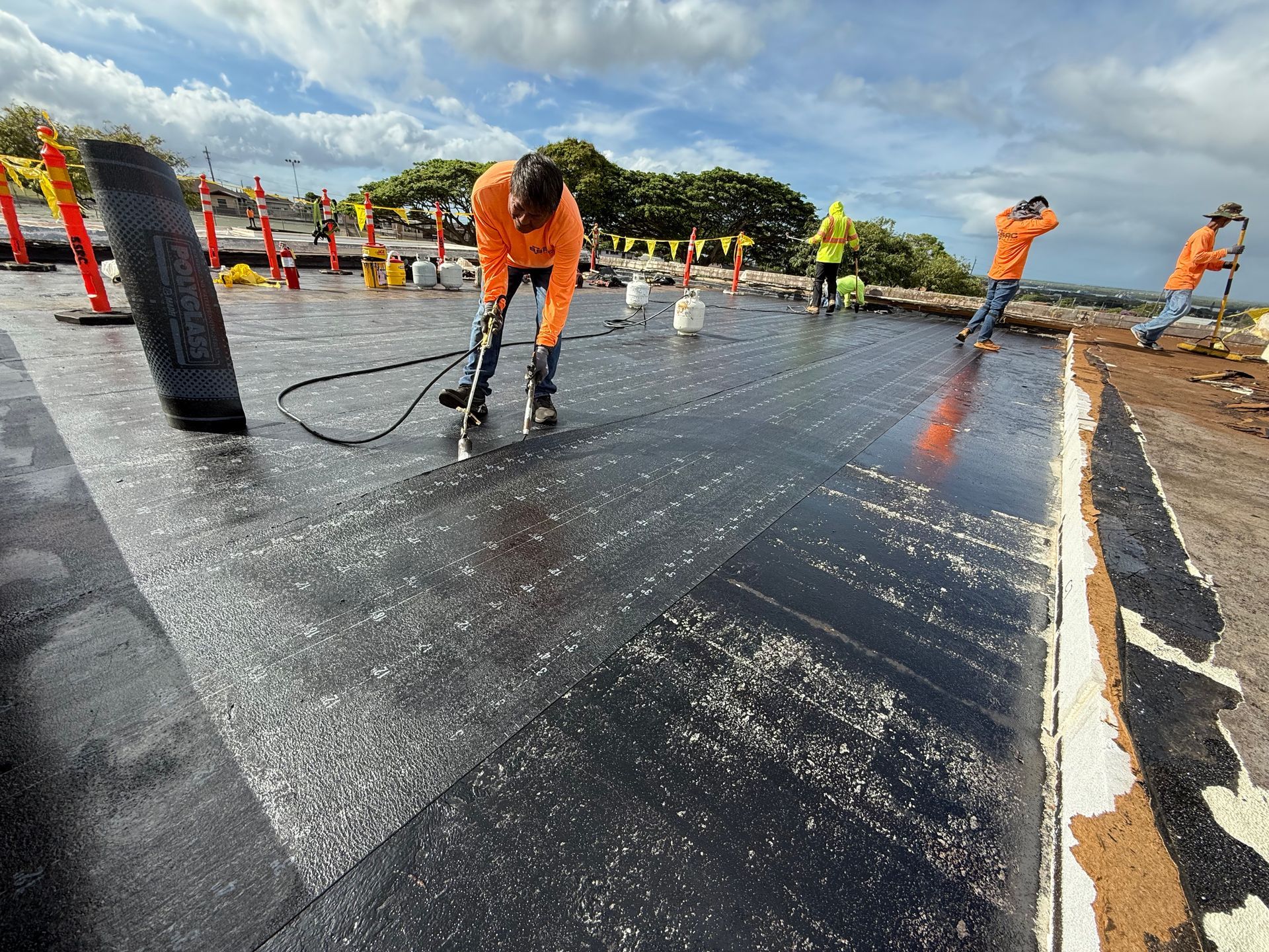 Construction workers installing roofing material on a flat rooftop, with tools and equipment visible.