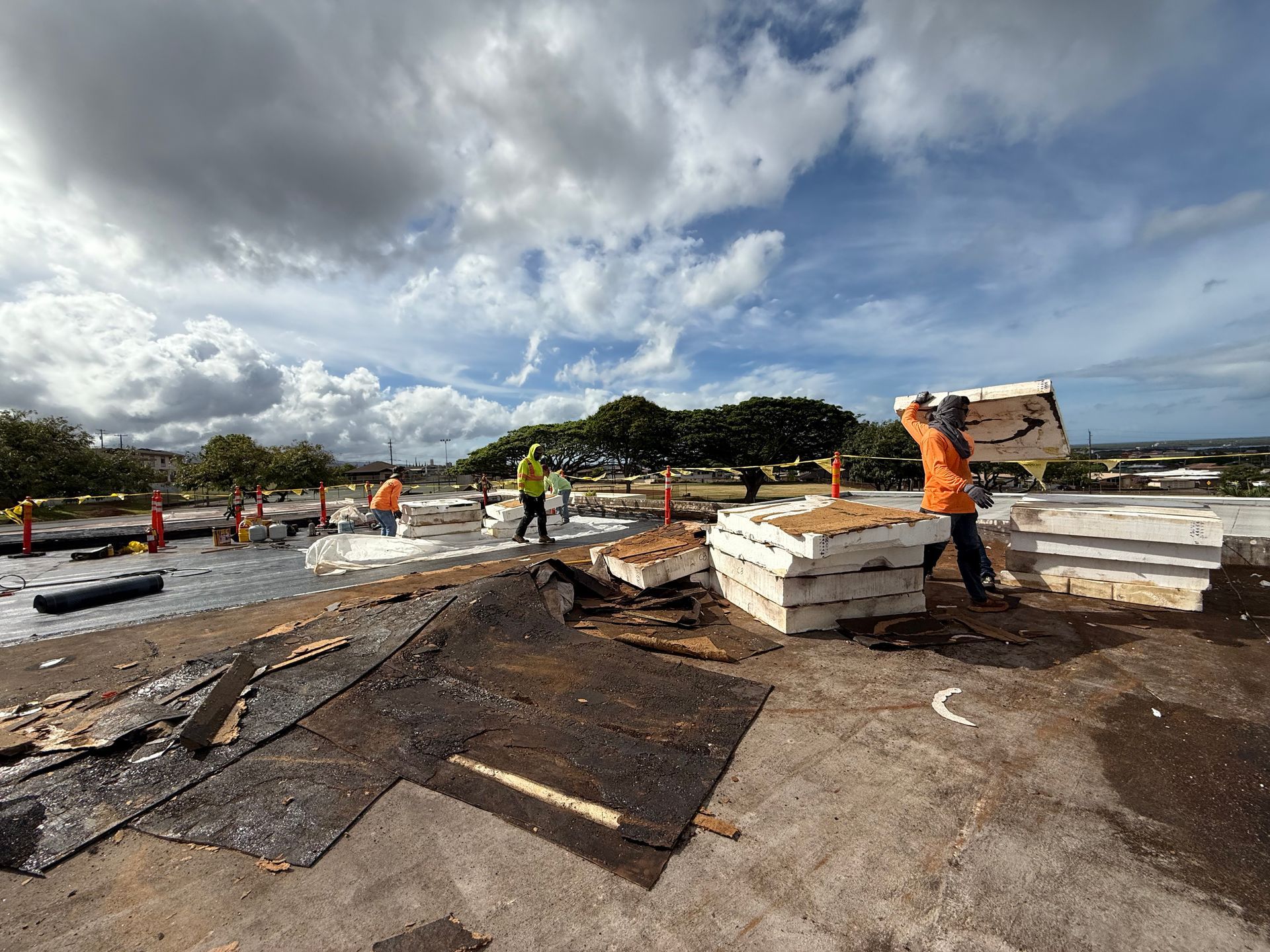 Construction workers on a roof remove old materials, carrying foam insulation against a cloudy sky.