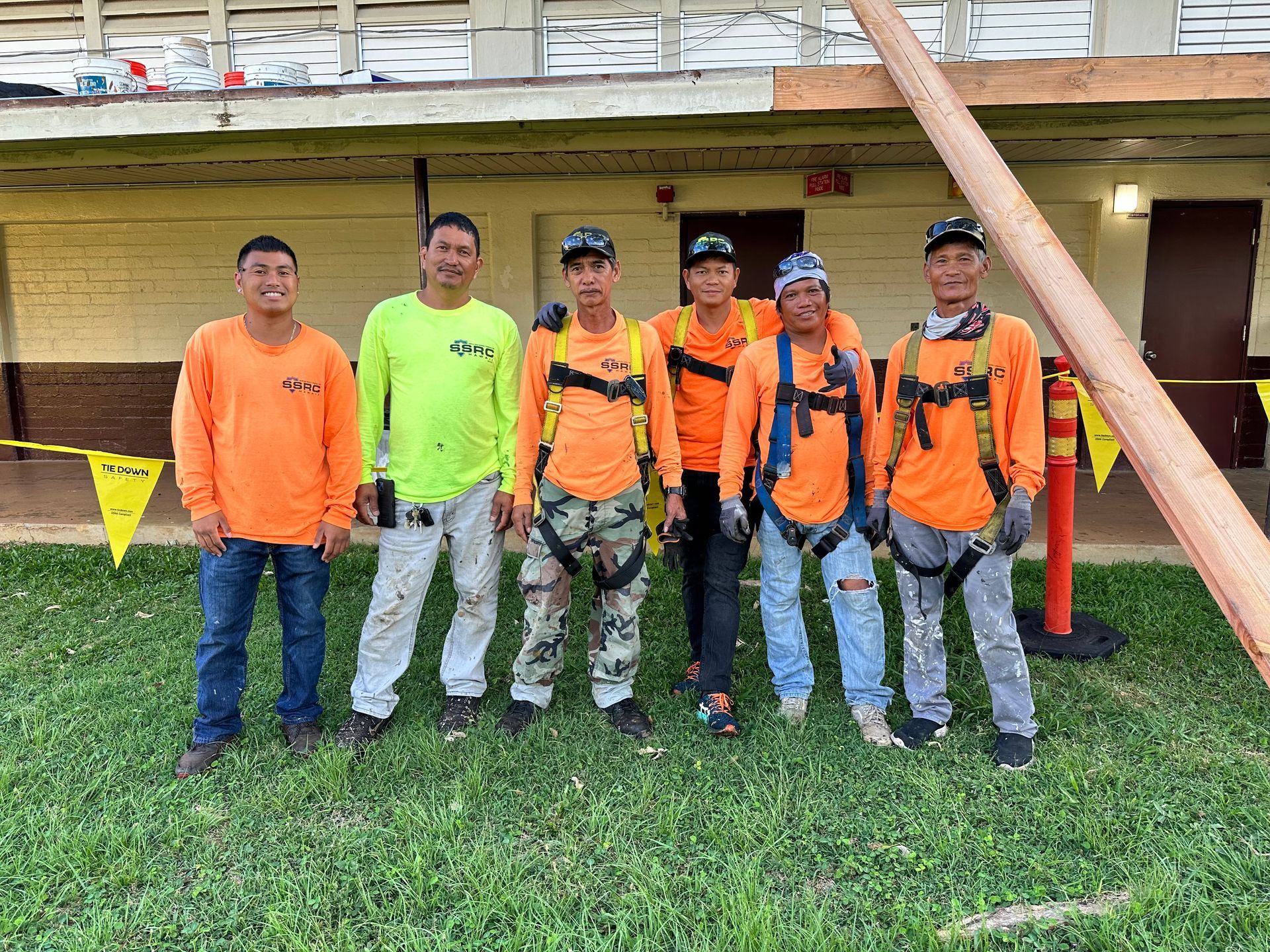 Group of construction workers in orange shirts and safety harnesses posing outdoors with a utility pole near a building.