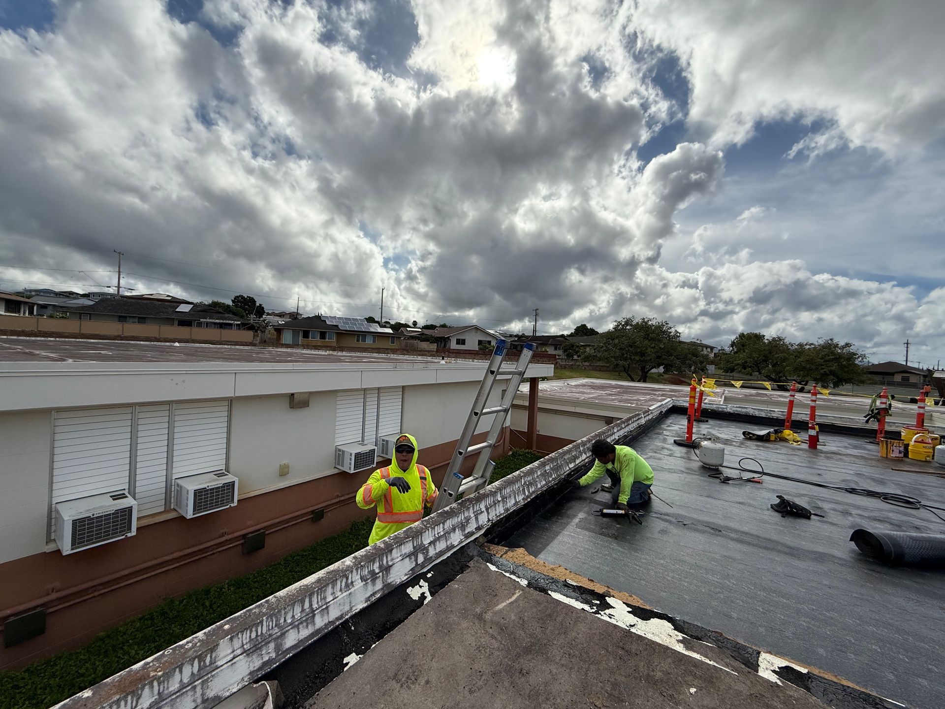 Two workers on a flat roof, repairing and inspecting. Cloudy sky overhead, building in background.