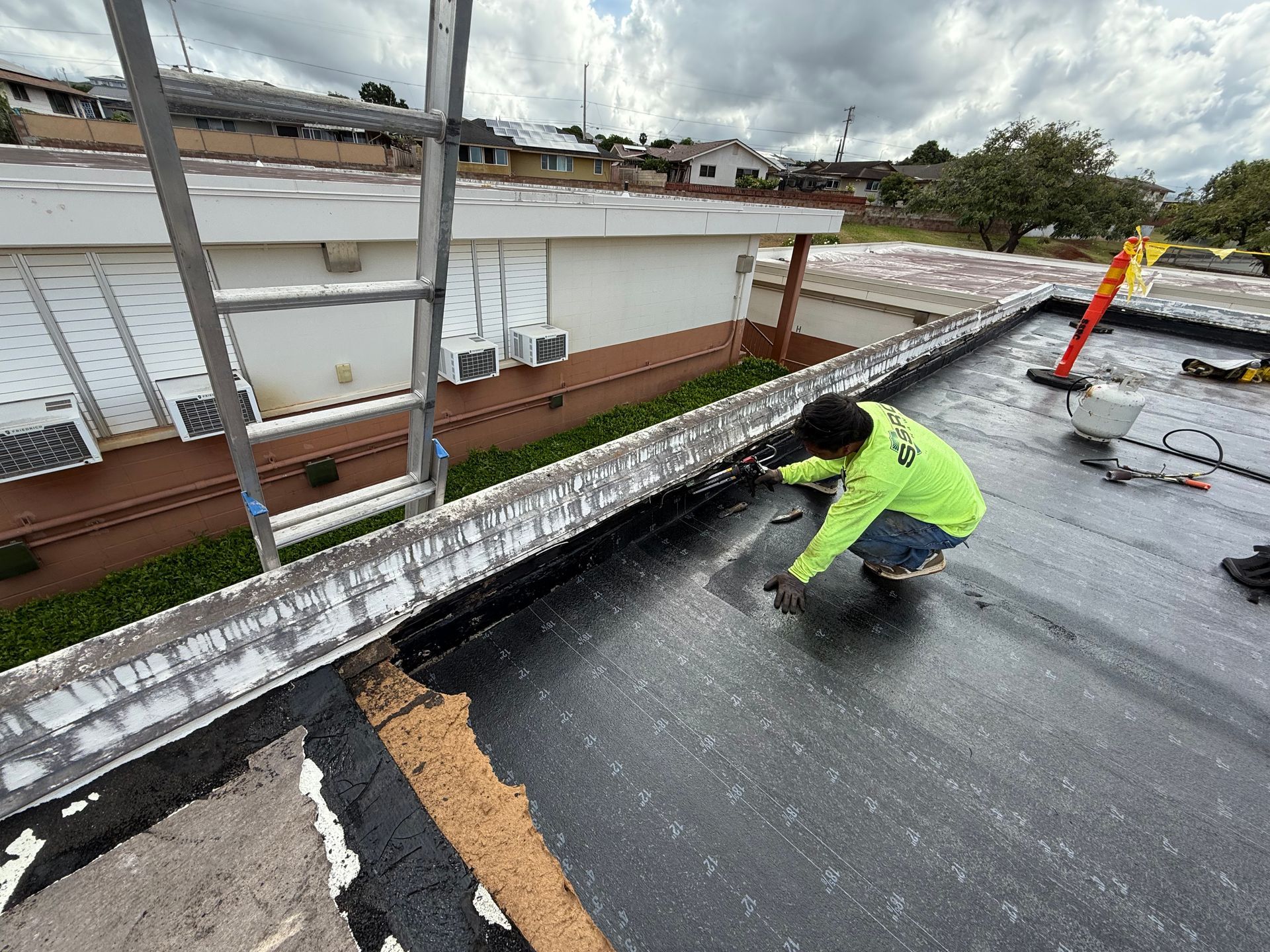 Person in neon shirt repairing flat roof with tools, ladder, and residential setting.