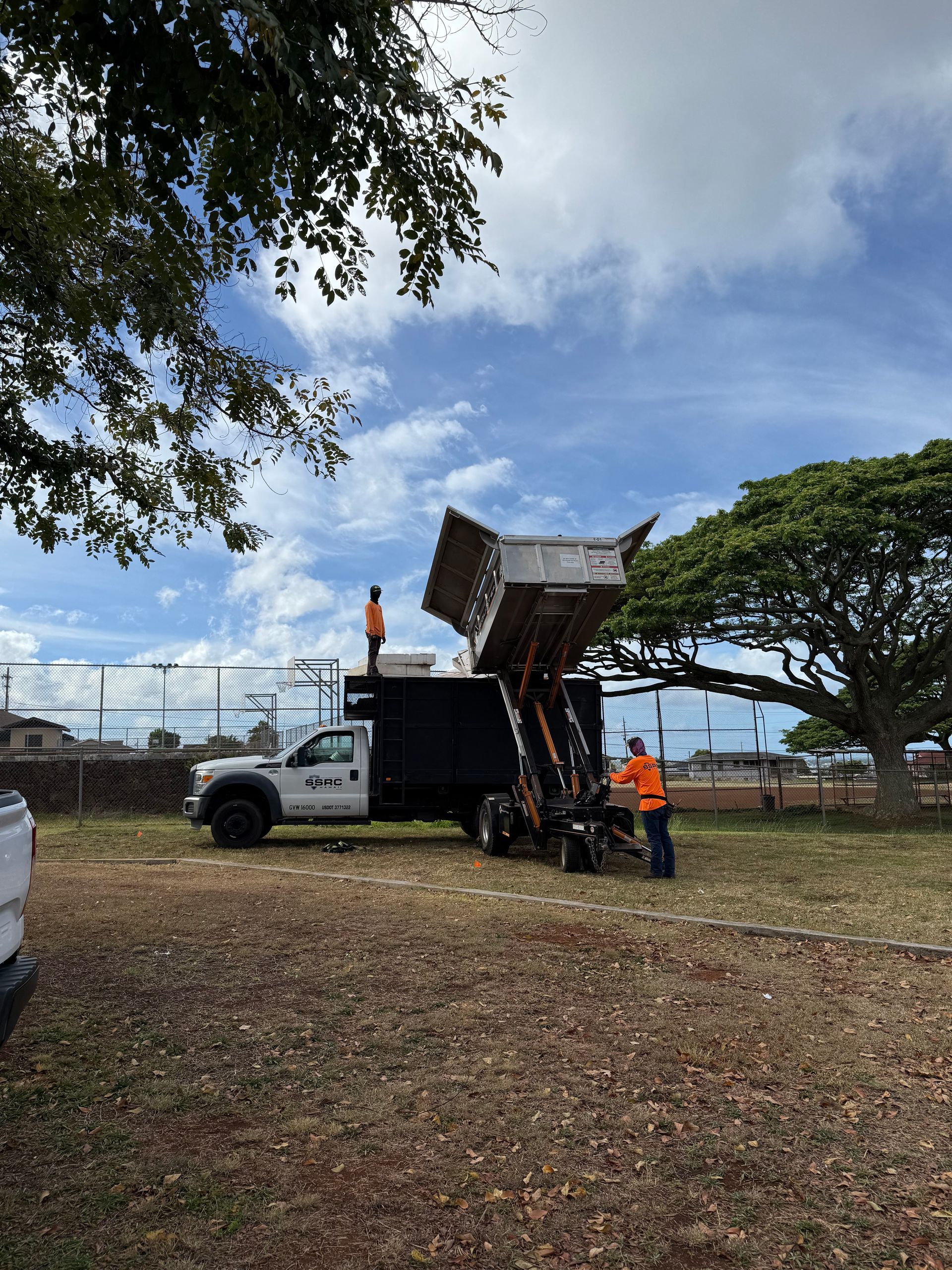 Workers emptying trash into a dump truck, sunny day. One worker stands on top. Black truck.
