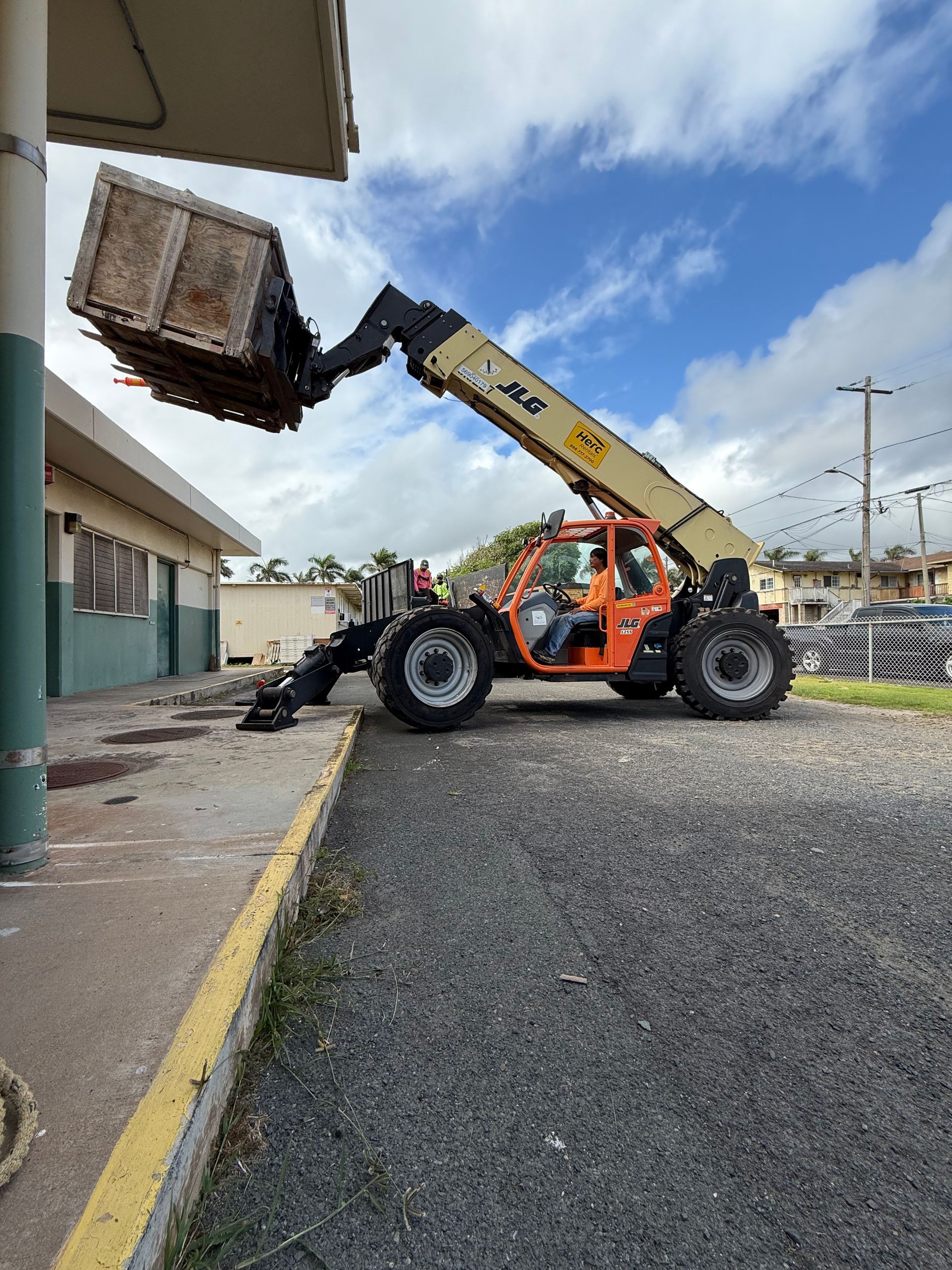 Orange JLG telehandler lifting wooden crate by a light green building on a gravel lot with blue sky.