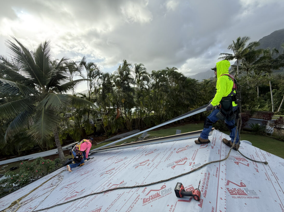 Two roofers in safety gear install roofing materials on a roof, tropical background.