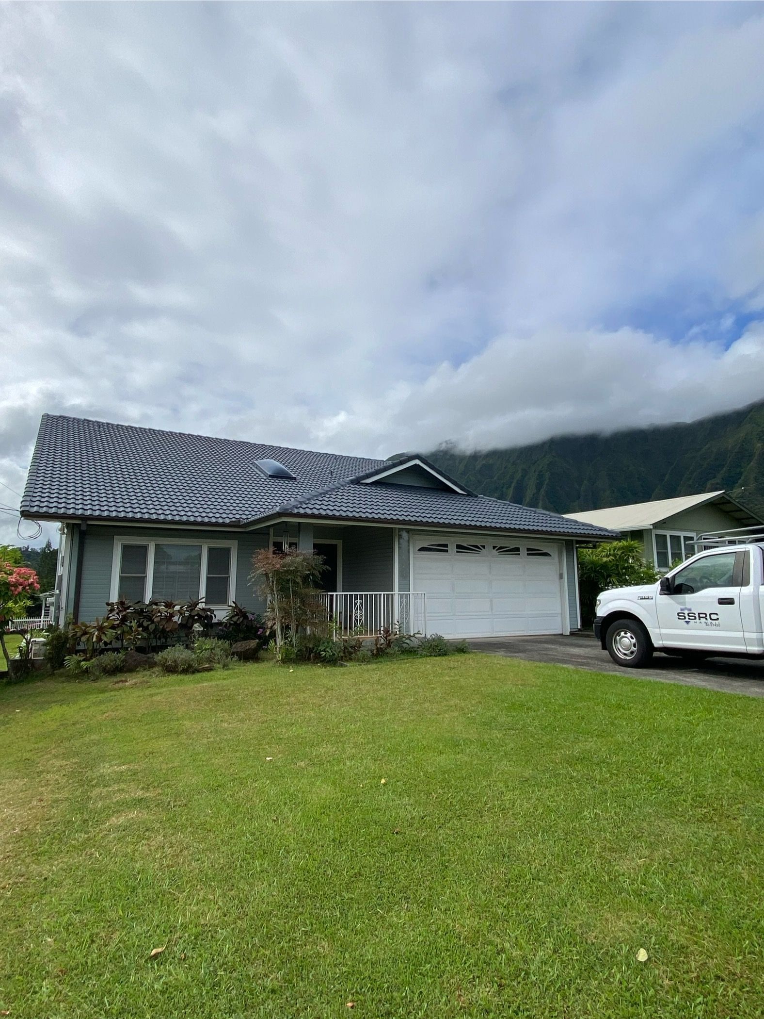 Gray house with a dark roof and white garage door, green lawn, cloudy sky, and a white truck.