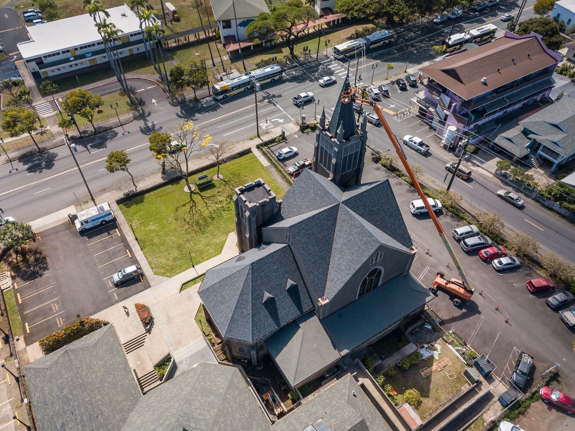 Aerial view of a gray-roofed church with a tall steeple, surrounded by roads, cars, and buildings in an urban setting.