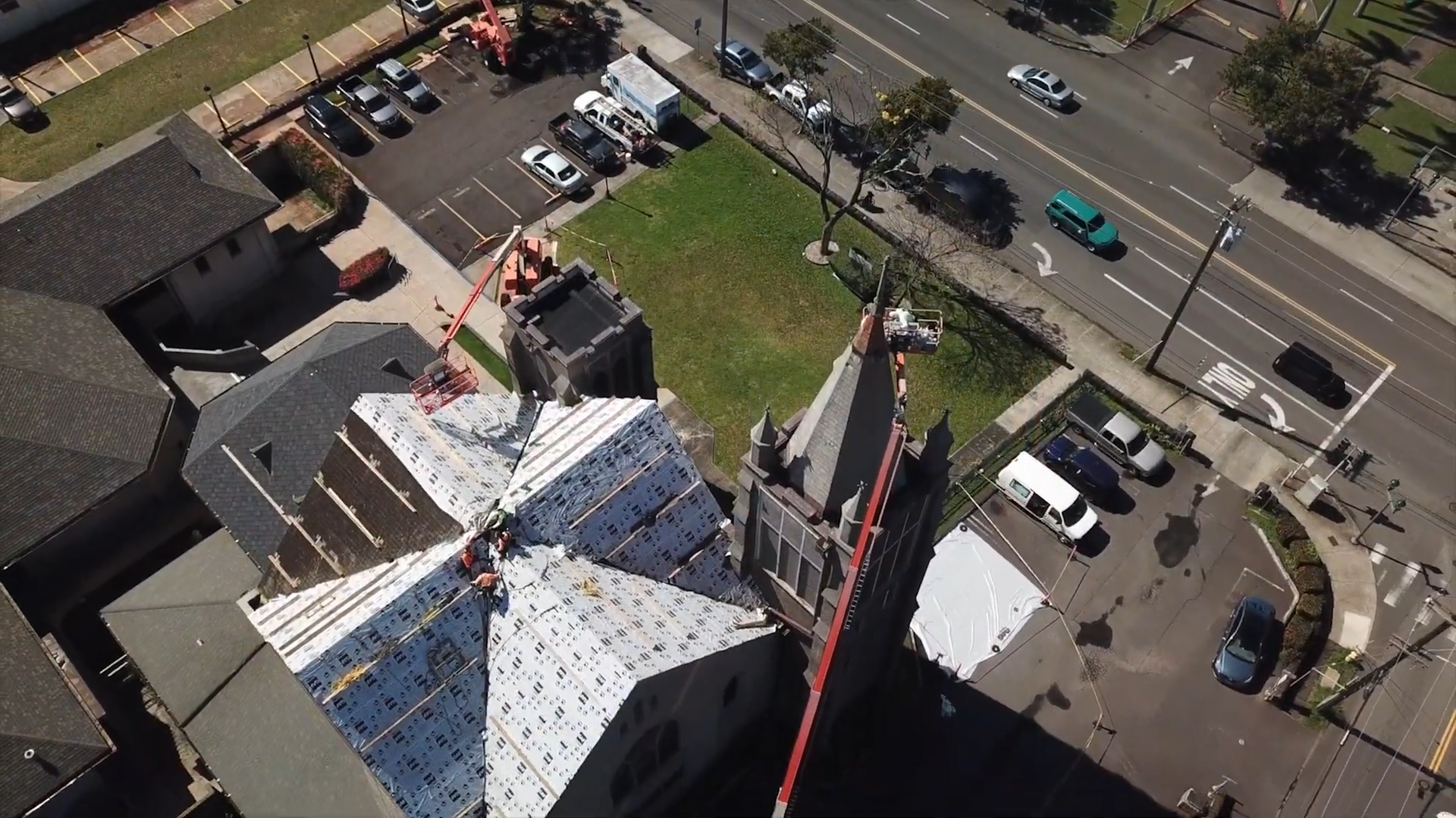 Aerial view of a church with construction on the roof, near a road with traffic and parking.