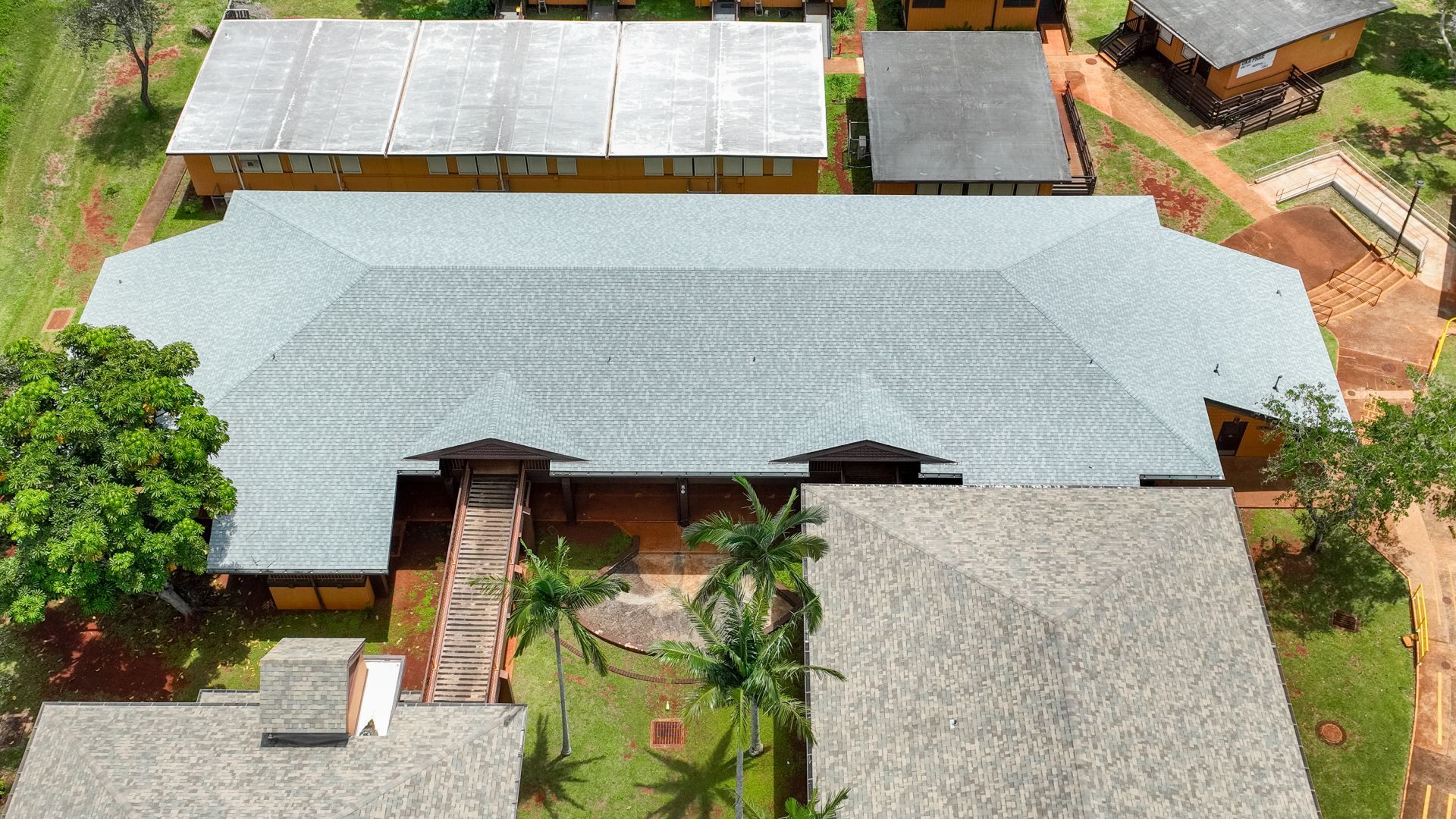 Aerial view of a gray-roofed building surrounded by other structures and green foliage.