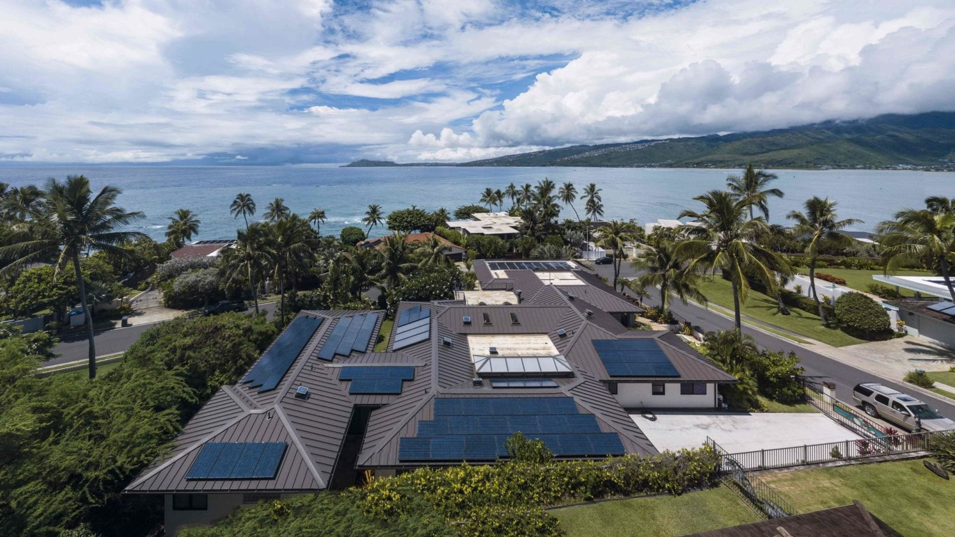 Aerial view of a house with solar panels on its roof, near a beach with ocean and mountains.