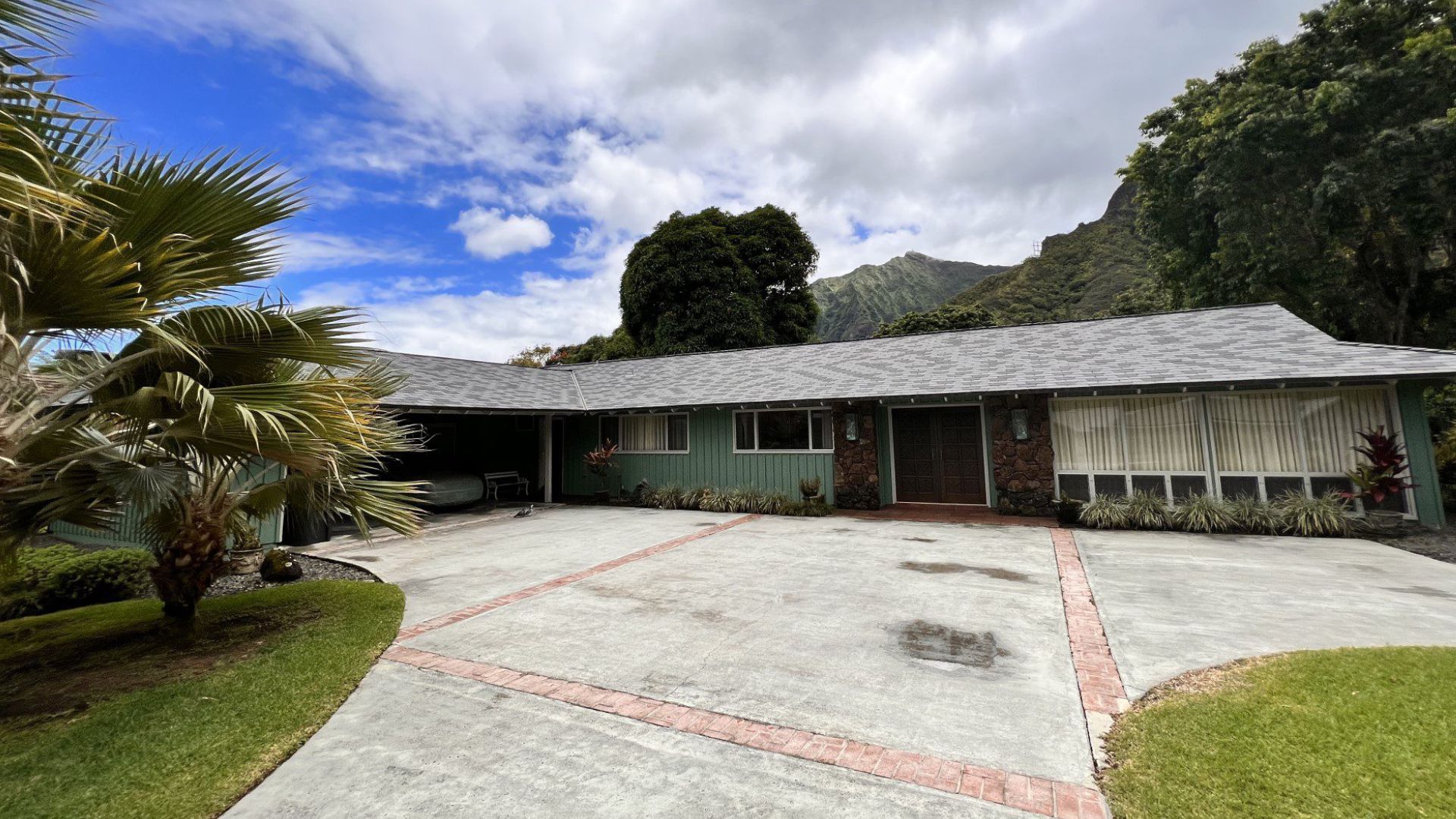House with green siding and a concrete driveway, nestled in lush greenery under a cloudy sky.